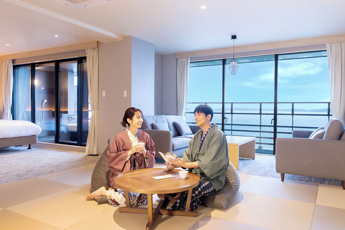 Une chambre d’hôtel moderne dotée d’une grande porte-fenêtre coulissante donnant sur un balcon avec vue sur la mer. On y trouve un lit, un canapé et une table ronde autour de laquelle sont assis deux personnes.