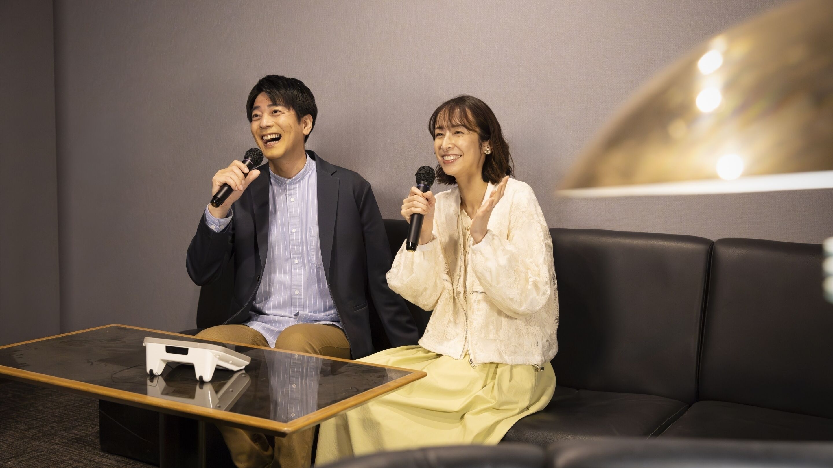 Two individuals sitting on a black leather couch with microphones, a tablet on a glass table, and a lamp in the background.