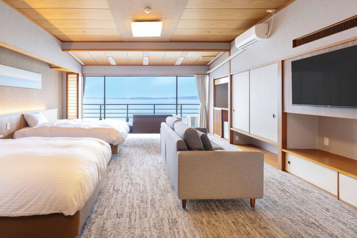Une chambre d’hôtel moderne équipée de deux lits, d’un canapé, d’une télévision et d’une grande fenêtre donnant sur la mer.