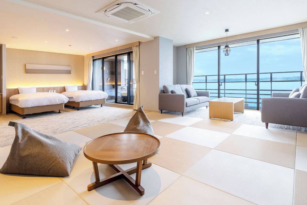Une chambre d’hôtel moderne avec balcon, deux lits, un canapé, une table basse et vue sur la mer.