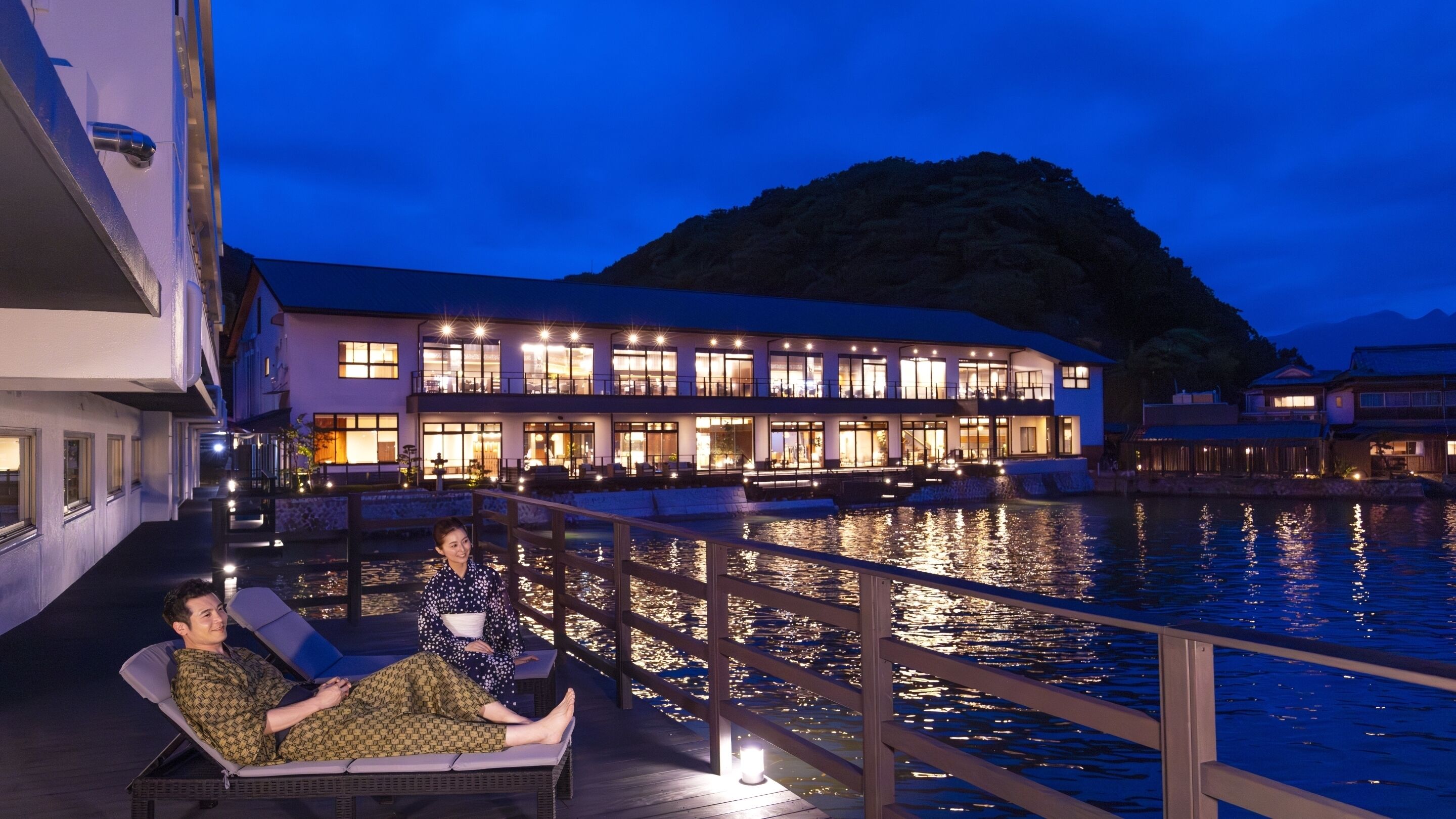 A waterfront property with a large building featuring multiple balconies and a pool area with lounge chairs.