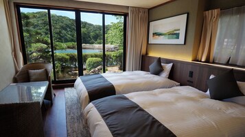 Habitación de hotel con dos camas, vista a un bosque y al agua, un comedor con mesa y sillas, y un cuadro en la pared.