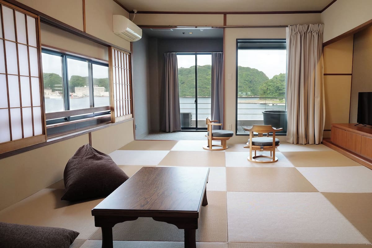 Una habitación japonesa tradicional con suelo de tatami, una mesita baja, dos sillas, un sofá, un televisor y vistas a un lago.