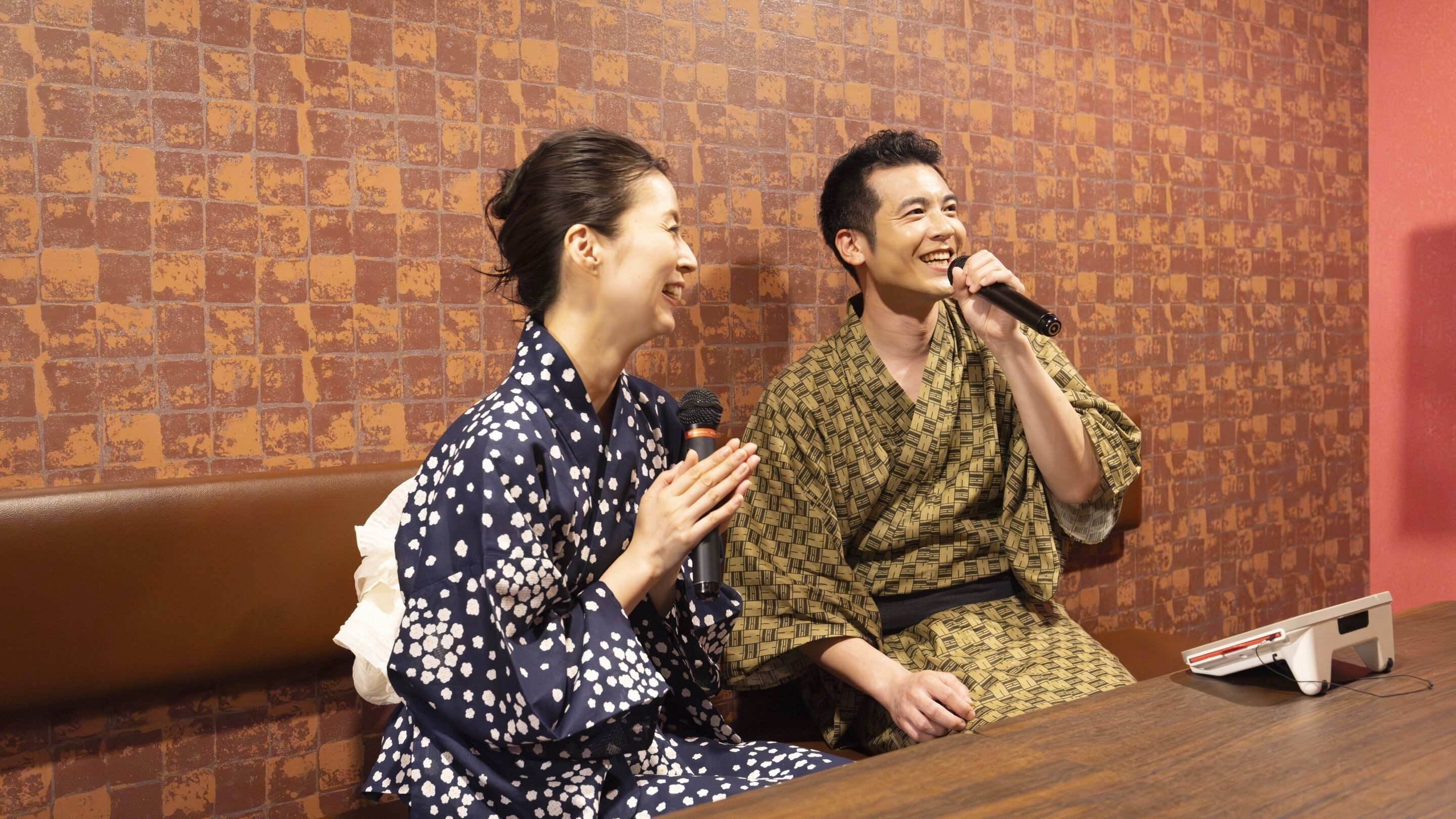 Two people sitting at a table with a microphone, in front of a patterned wall.