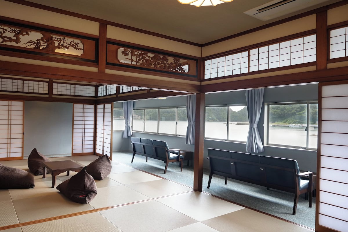 Una habitación tradicional japonesa con suelo de tatami, una mesa baja y cojines. Amplias ventanas ofrecen vistas a una masa de agua.
