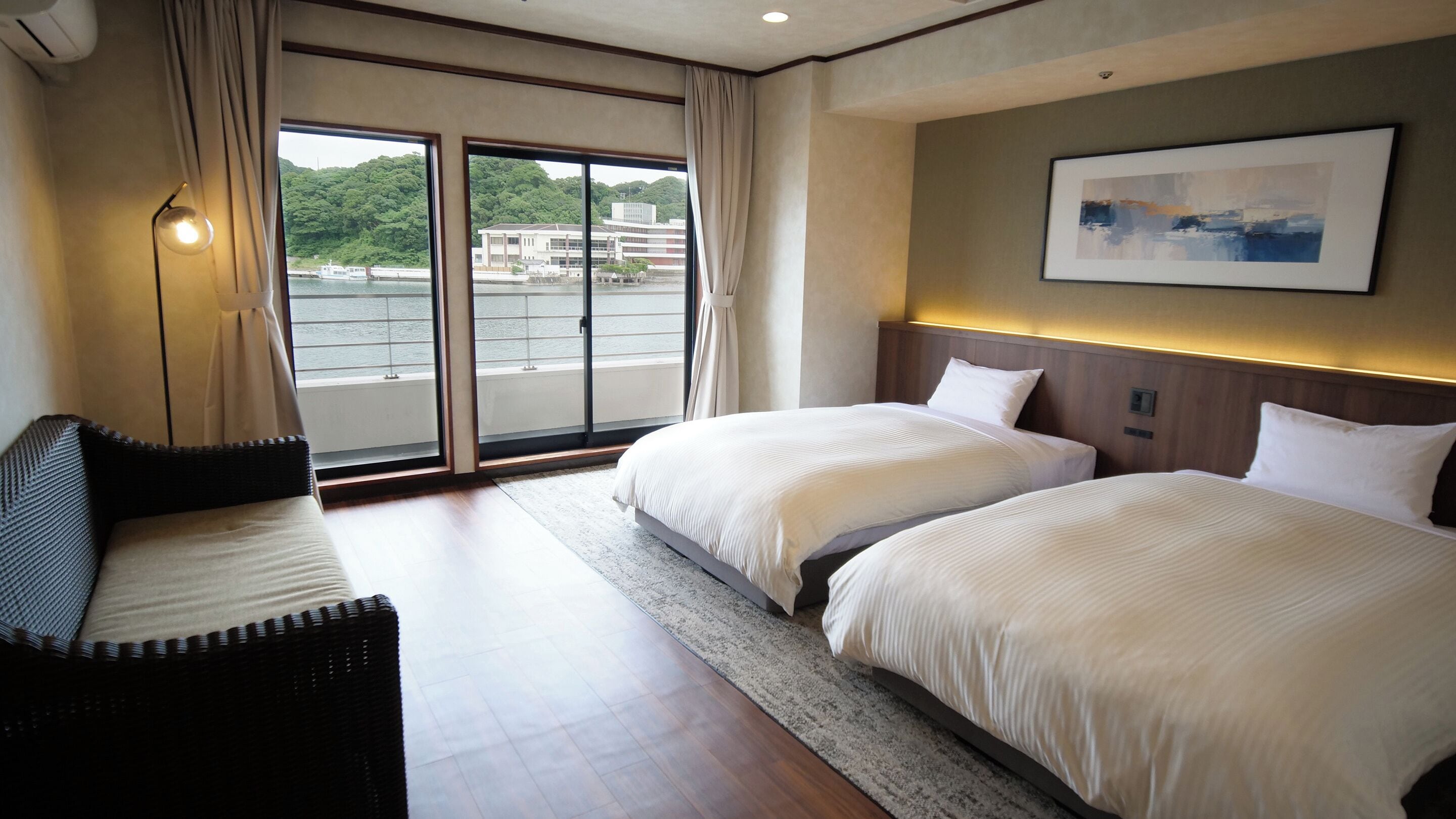 Habitación de hotel con dos camas, un sofá, un ventanal con vistas a edificios y follaje, y un cuadro en la pared.