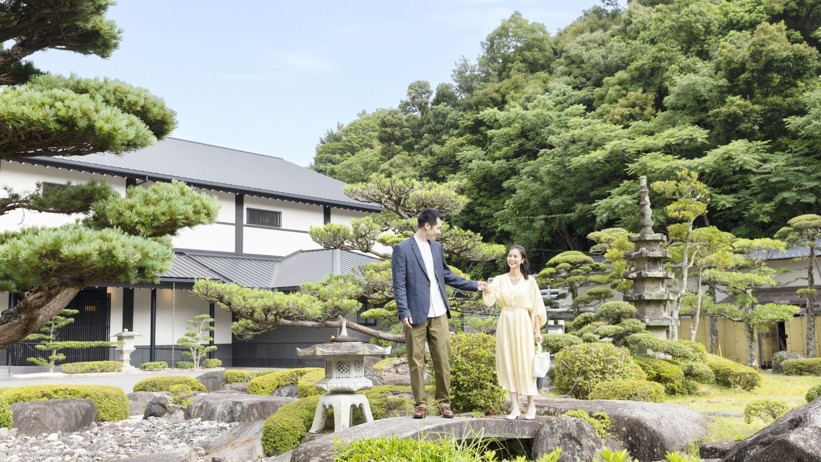 Ein traditioneller japanischer Garten mit einer Steinlaterne, einem Gebäude mit Ziegeldach und sorgfältig gestutzten Bäumen.