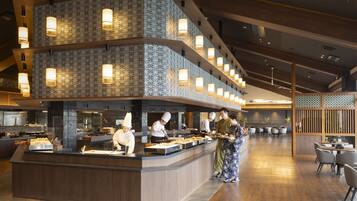 A modern restaurant with a central counter, chefs preparing food, and a seating area with tables and chairs.