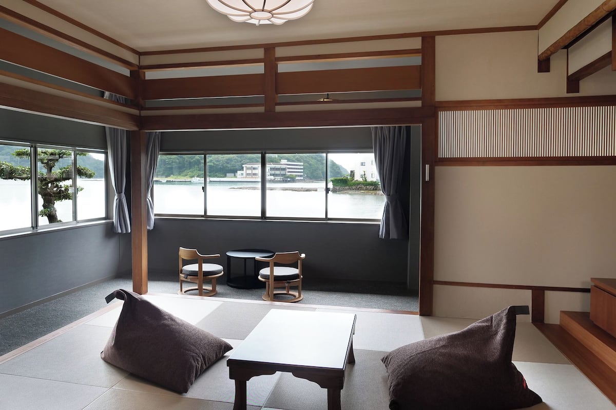 Una habitación con esteras de tatami, una mesa baja, dos sillas y vistas a un cuerpo de agua y edificios.