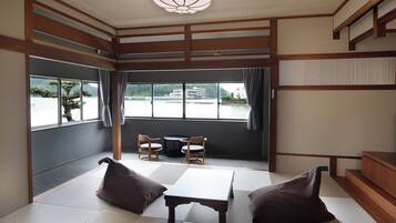 Ein Zimmer mit Tatami-Matten, einem niedrigen Tisch, zwei Stühlen und Blick auf ein Gewässer sowie Gebäude.