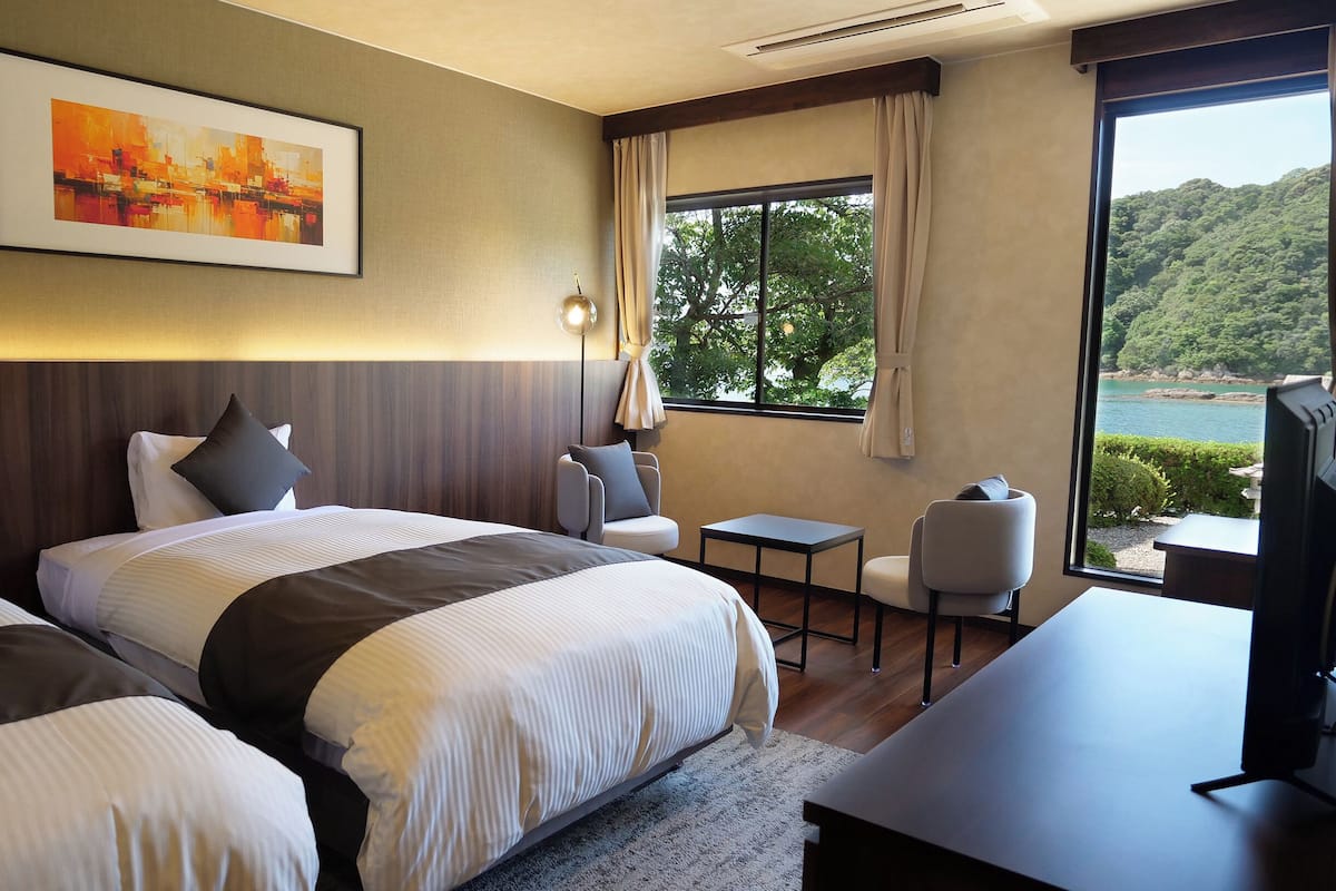 Habitación de hotel con dos camas, un escritorio, una silla, un televisor y un ventanal con vistas panorámicas.