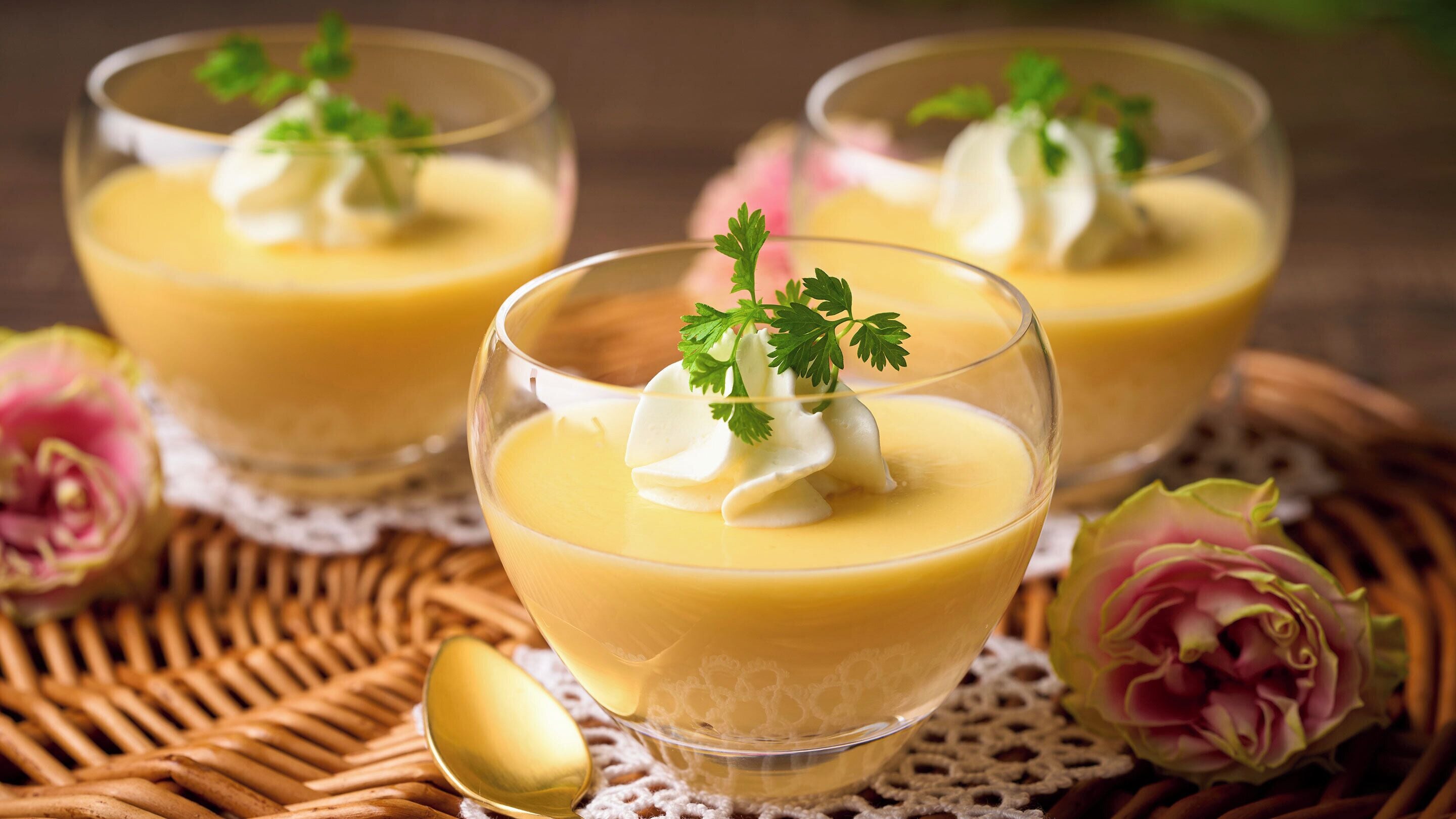 Three glasses of yellow dessert with whipped cream and parsley garnish, placed on a woven mat with lace doily, surrounded by pink flowers.