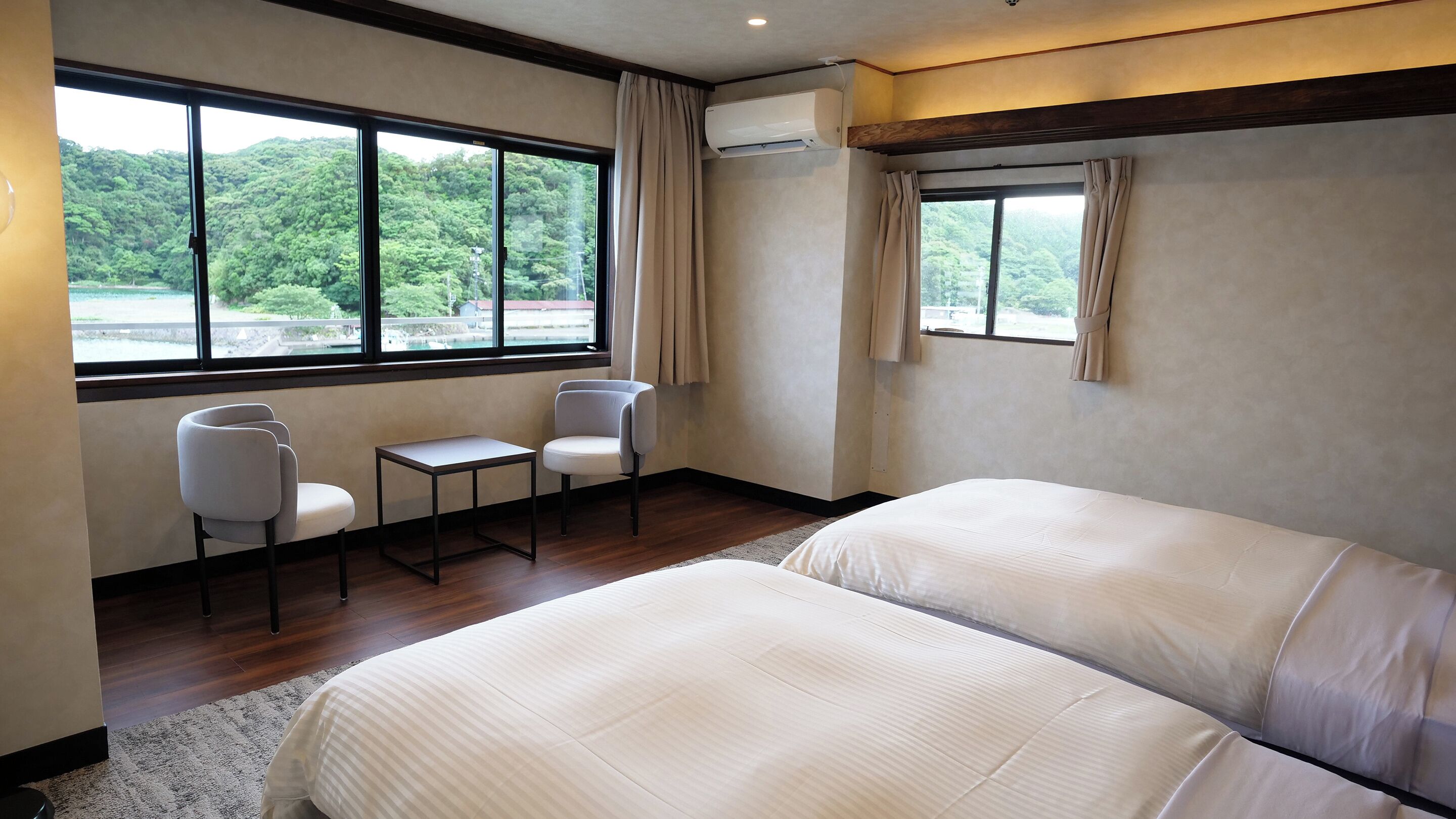 Ein Hotelzimmer mit zwei Betten, einem kleinen Tisch, zwei Stühlen und großen Fenstern mit Blick auf eine bewaldete Landschaft.