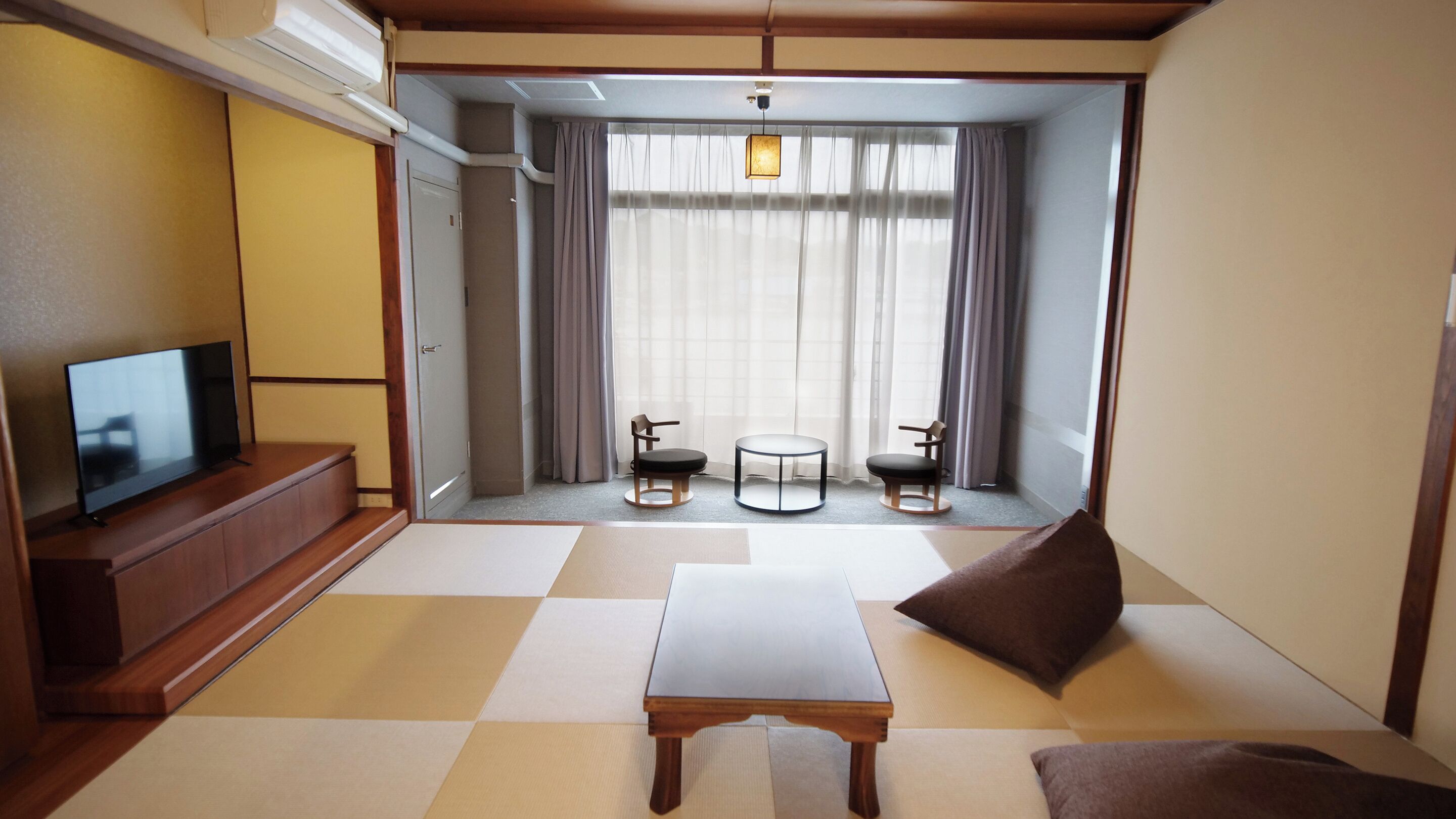 Quarto com televisão sobre suporte de madeira, uma mesinha, duas cadeiras e vista através de uma janela grande com cortinas.