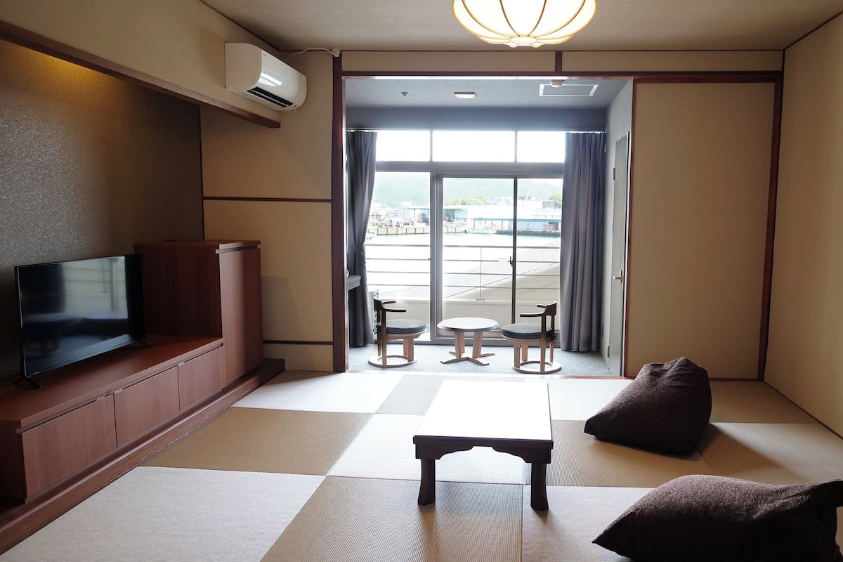 Habitación de hotel con suelo de tatami, televisor sobre un soporte de madera, una mesita y sillas, y vista a un estacionamiento a través de la puerta.