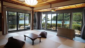 Una habitación con esteras de tatami, una mesa de madera y una silla de mimbre. Amplias ventanas ofrecen vistas a un cuerpo de agua y follaje.