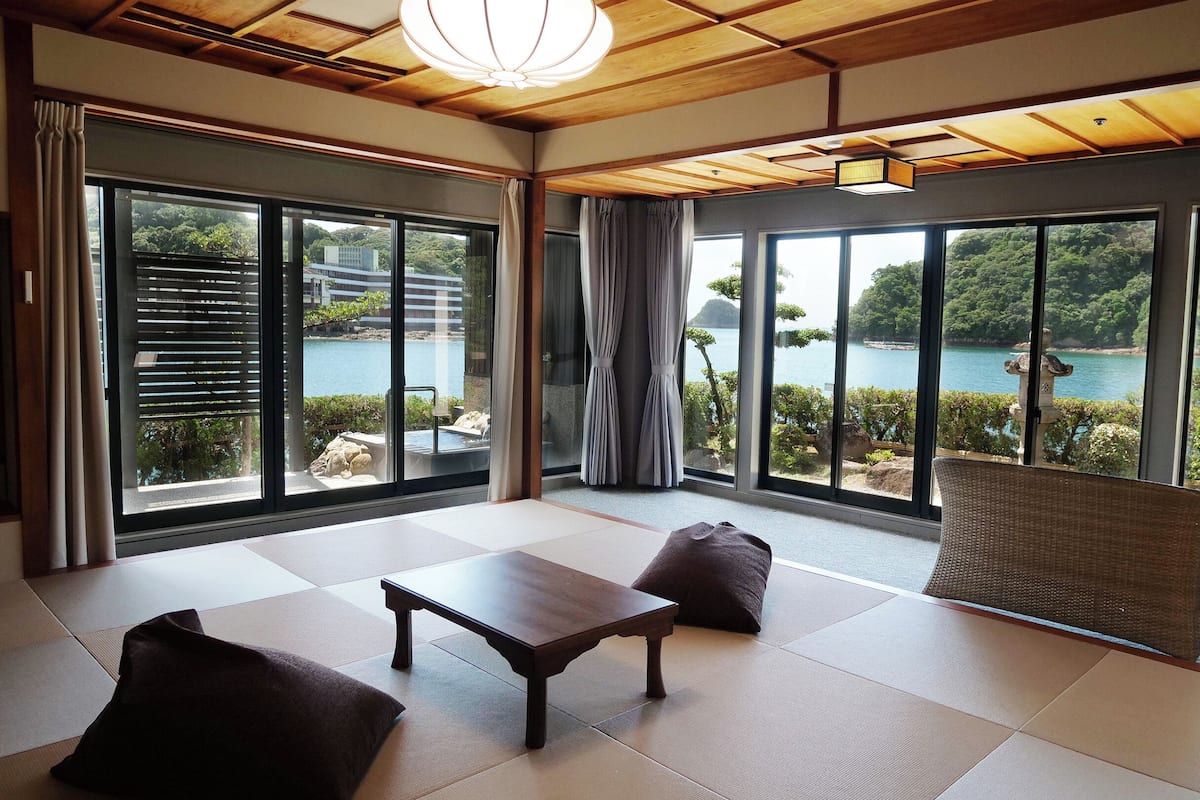 Una habitación con esteras de tatami, una mesa de madera y una silla de mimbre. Amplias ventanas ofrecen vistas a un cuerpo de agua y follaje.