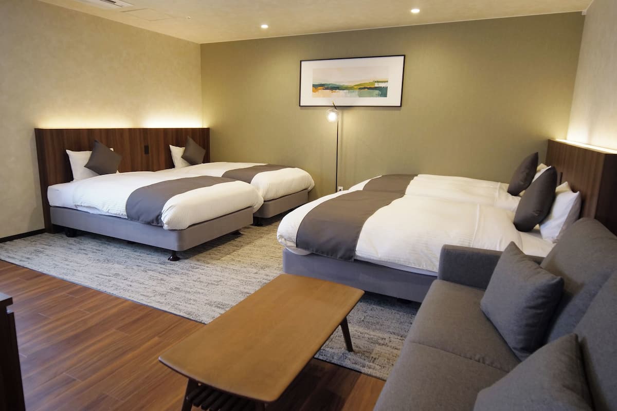 Habitación de hotel con dos camas, un sofá, una mesa de centro de madera y una lámpara de pie.