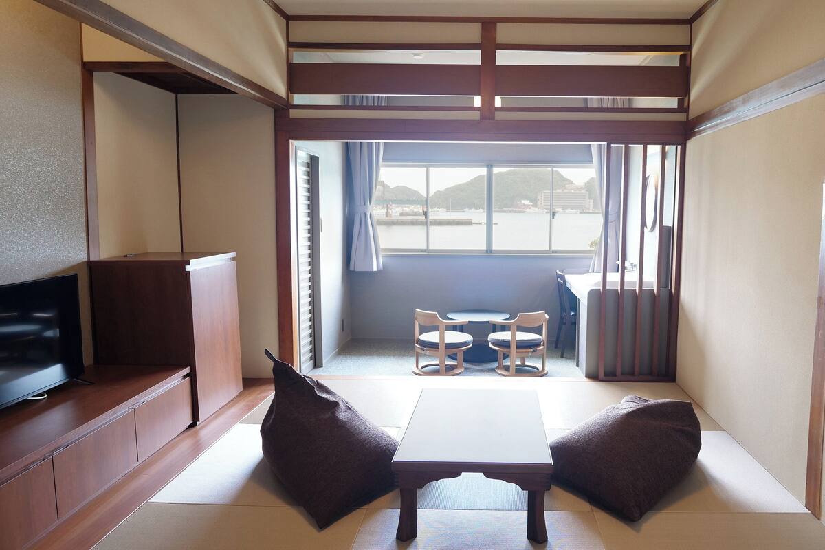 Una habitación con suelo de tatami, una mesa baja con dos sillas, un televisor y una puerta corrediza que da a vistas de un cuerpo de agua.