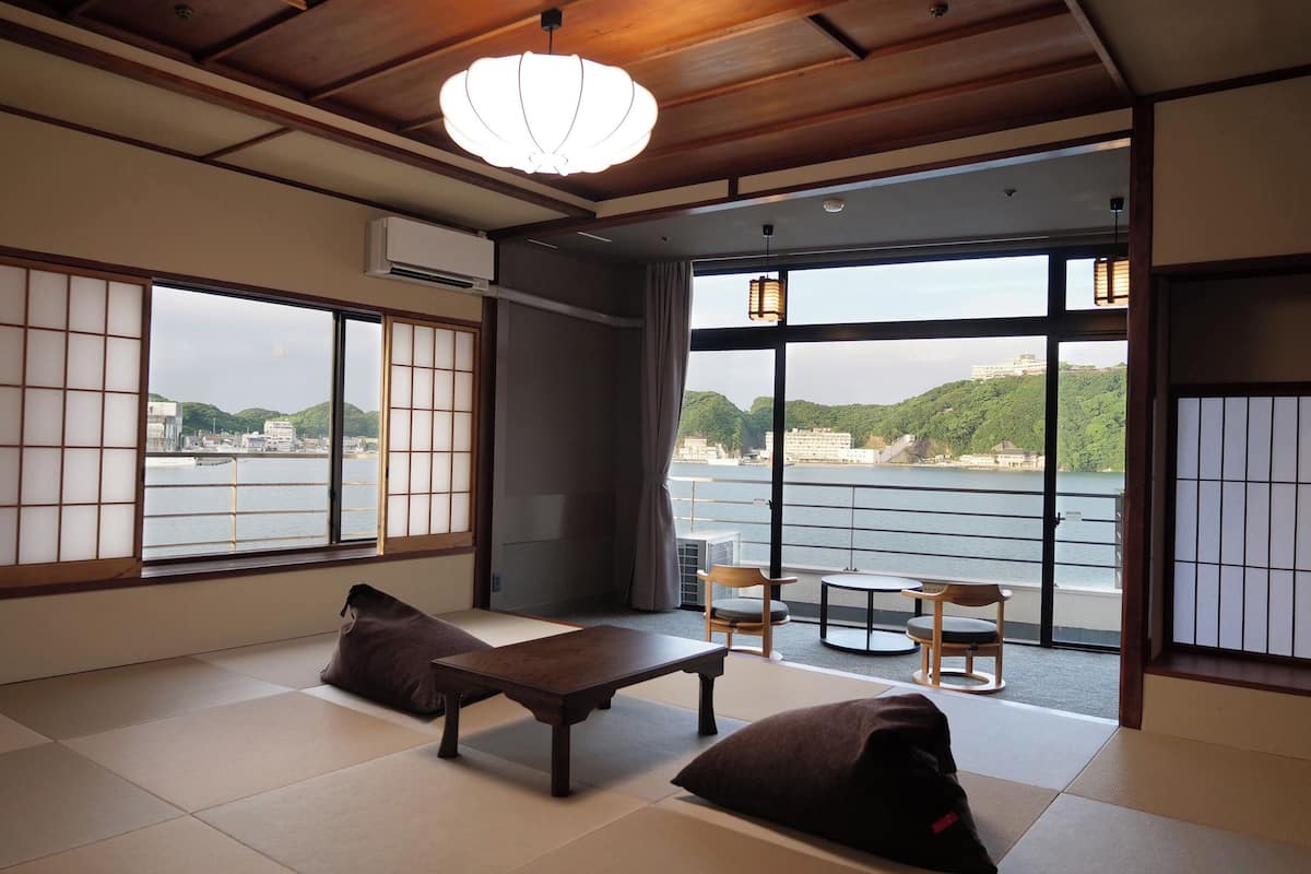 Una habitación tradicional japonesa con suelo de tatami, una mesa baja, dos cojines y grandes ventanales con vistas a un cuerpo de agua y un paisaje urbano.