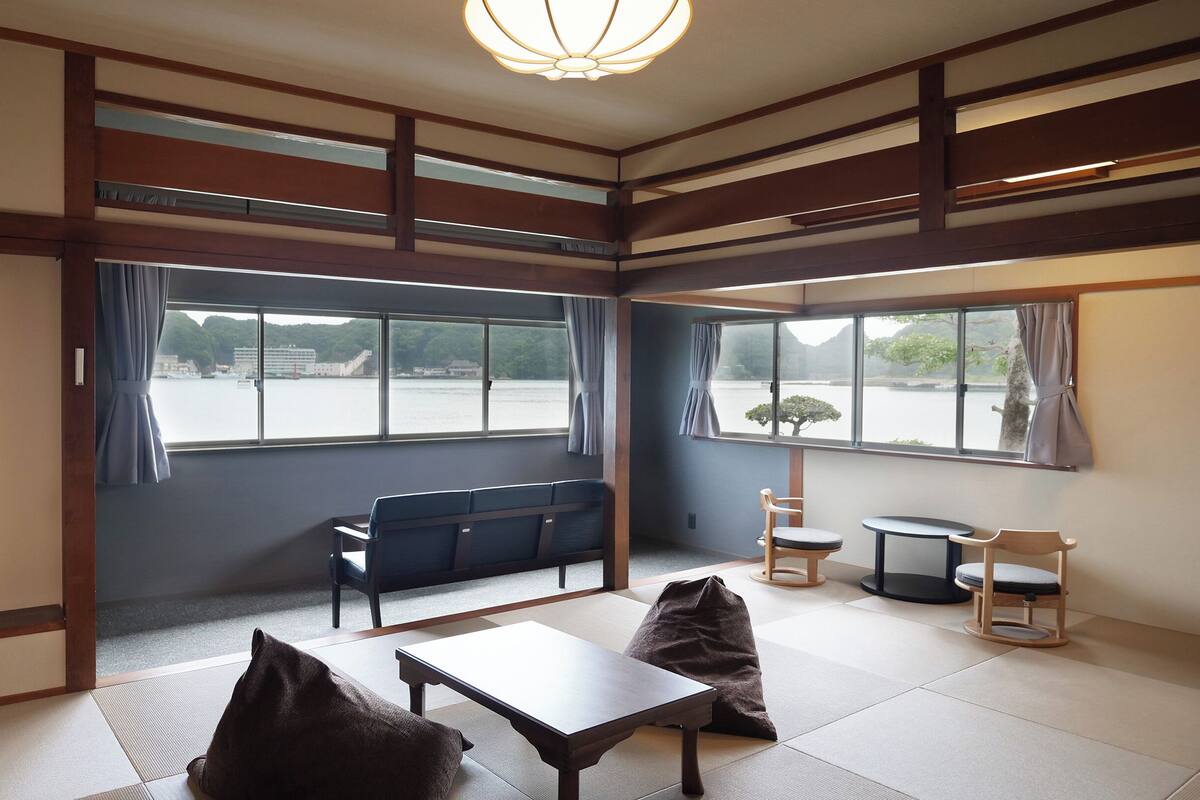 Una habitación tradicional japonesa con suelo de tatami, una mesa baja, un sofá y dos sillas. Amplias ventanas ofrecen vistas a una masa de agua y vegetación.
