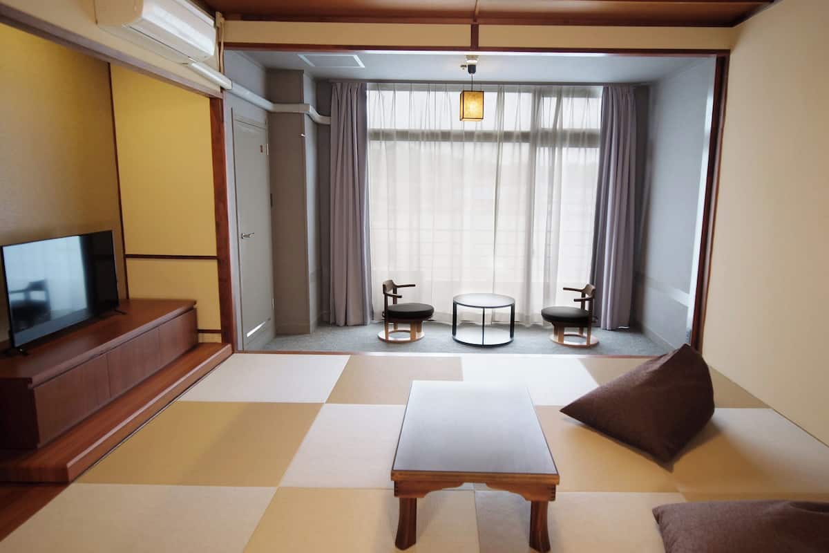 Una habitación con una televisión sobre un mueble de madera, una mesita, dos sillas y vistas a través de un ventanal con cortinas.