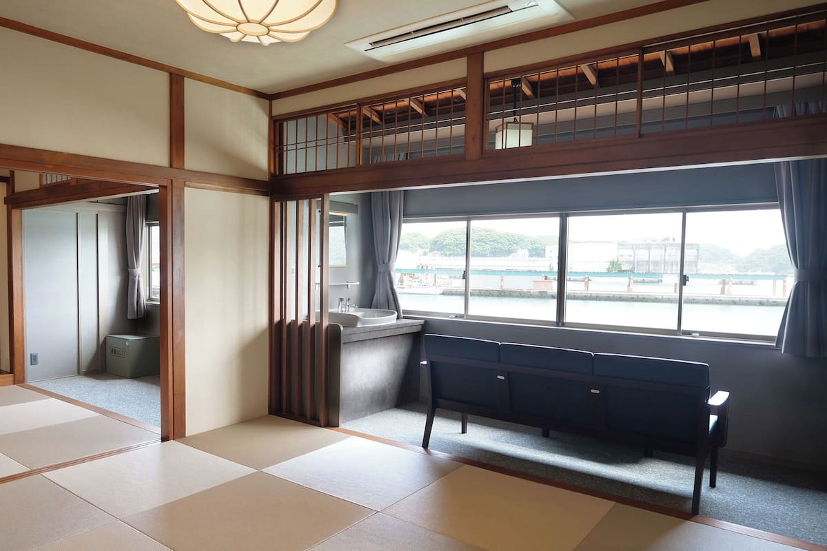 Una habitación japonesa tradicional con suelo de tatami, una mesa baja, un televisor y vista a un cuerpo de agua.