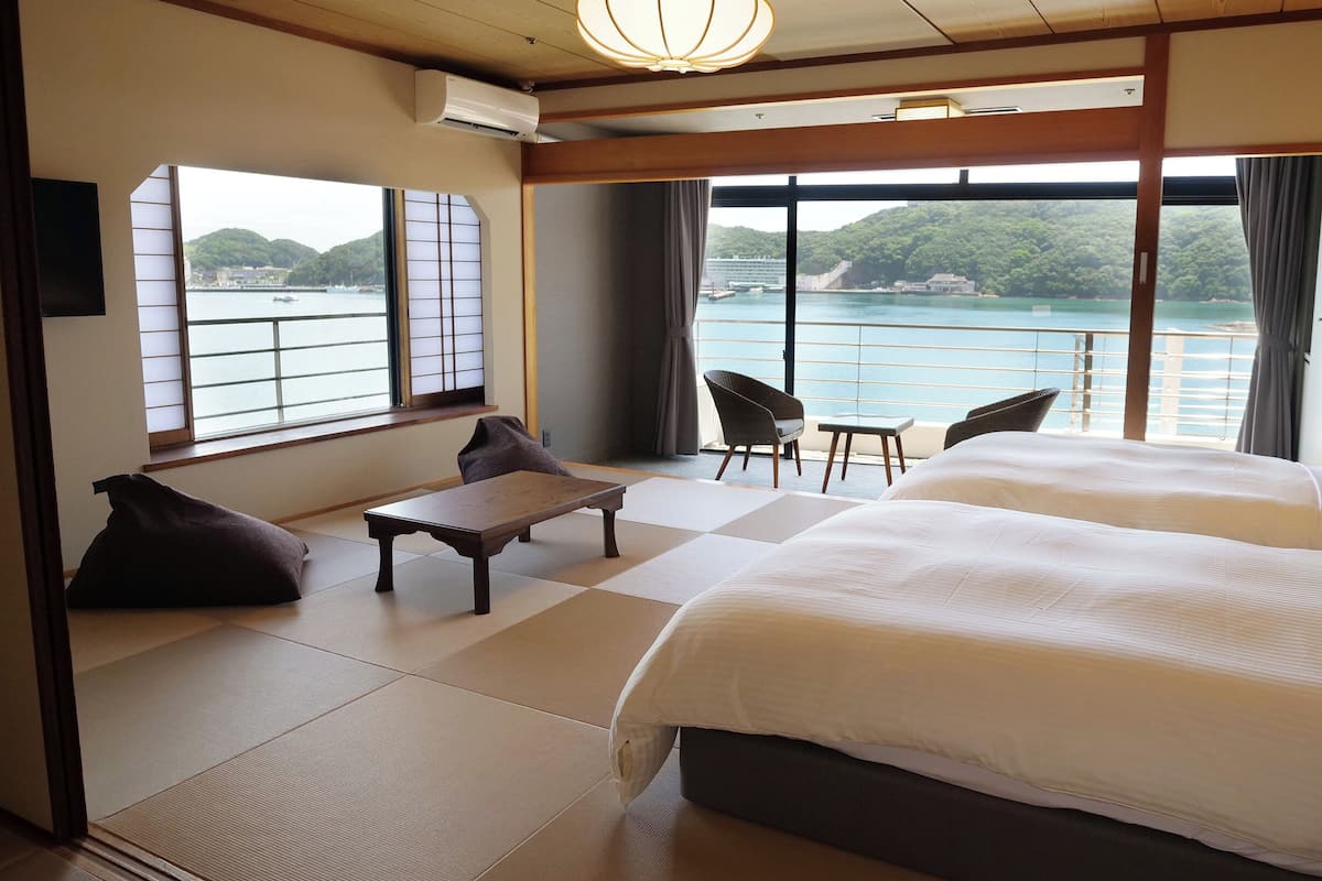 Habitación de hotel con dos camas, una mesita, sillas y ventanas amplias que dan a una masa de agua y vegetación.