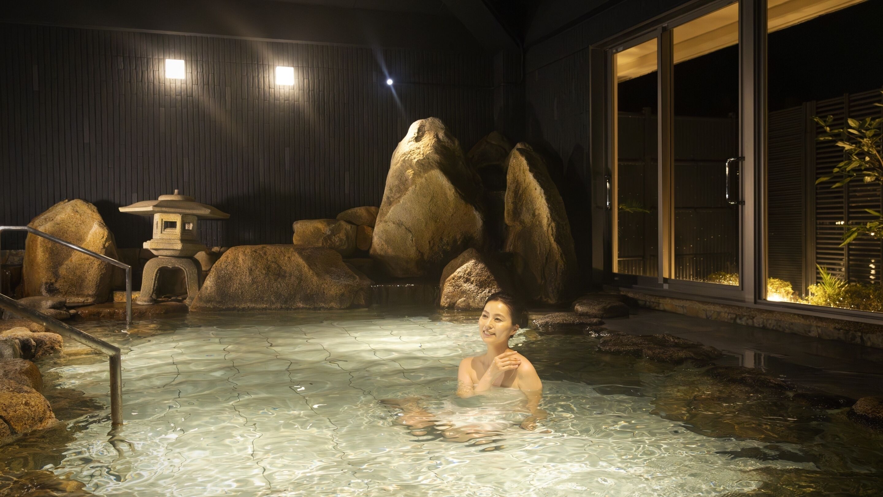 A person in a hot spring with large rocks and a pagoda.