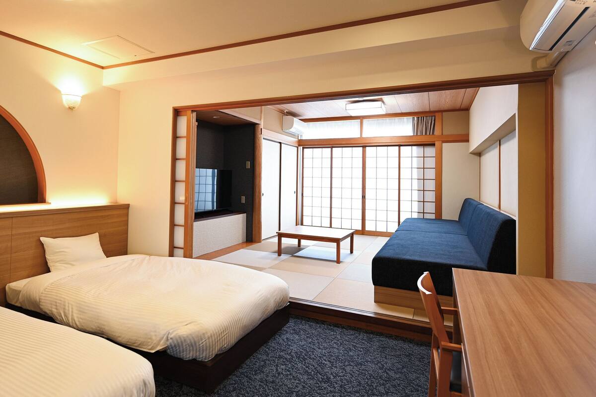 A hotel room with two beds, a sofa, a TV, and a sliding door leading to a balcony.