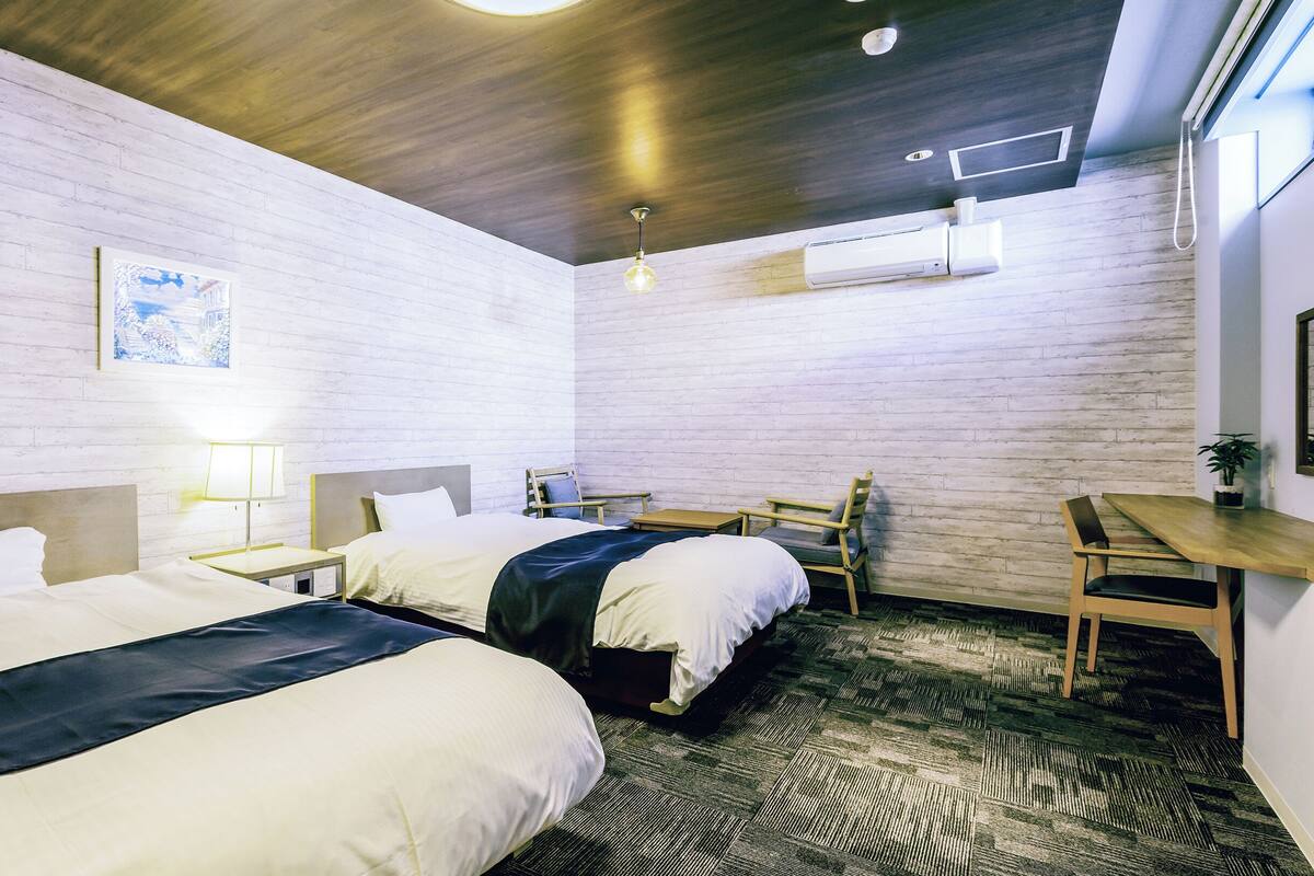 A hotel room with two beds, a desk, a chair, a lamp, and a ceiling fan.