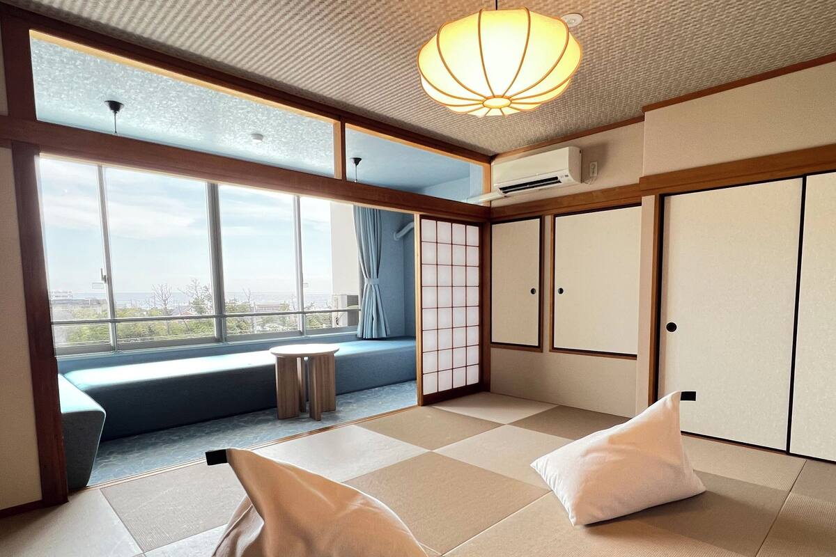 A room with tatami flooring, a sliding door, a view through a large window, and a ceiling light fixture.