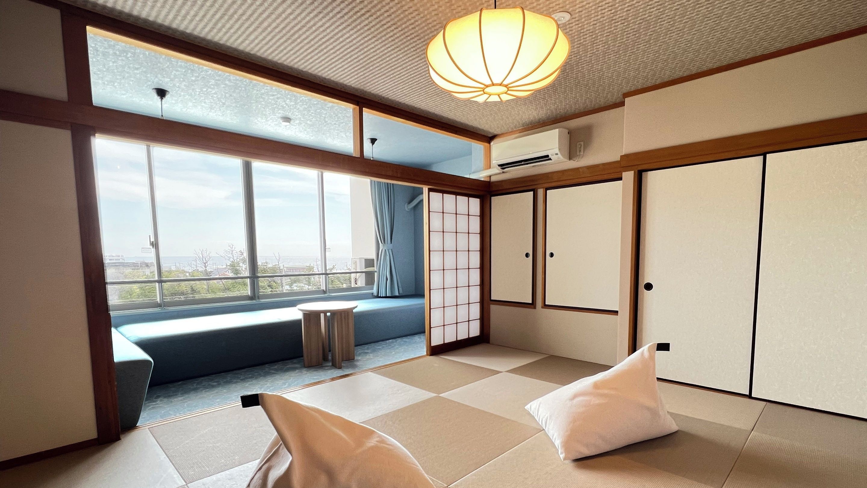Una habitación con suelo de tatami, una puerta corredera, vistas a través de un ventanal grande y una lámpara en el techo.
