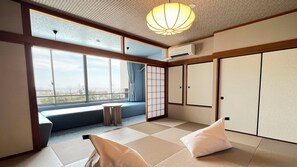 A room with tatami flooring, a sliding door, a view through a large window, and a ceiling light fixture.