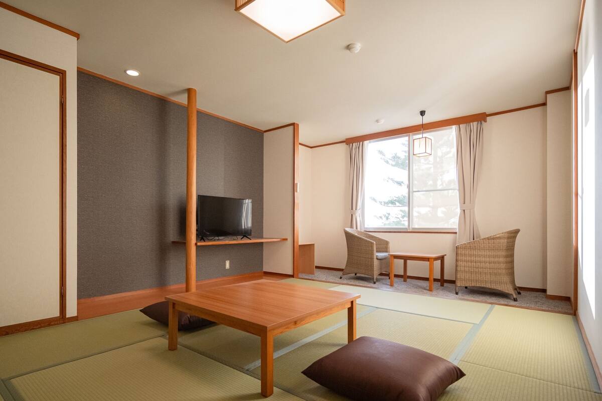 A traditional Japanese room with tatami flooring, a low wooden table, a television on a shelf, and a view of the outdoors through a window.