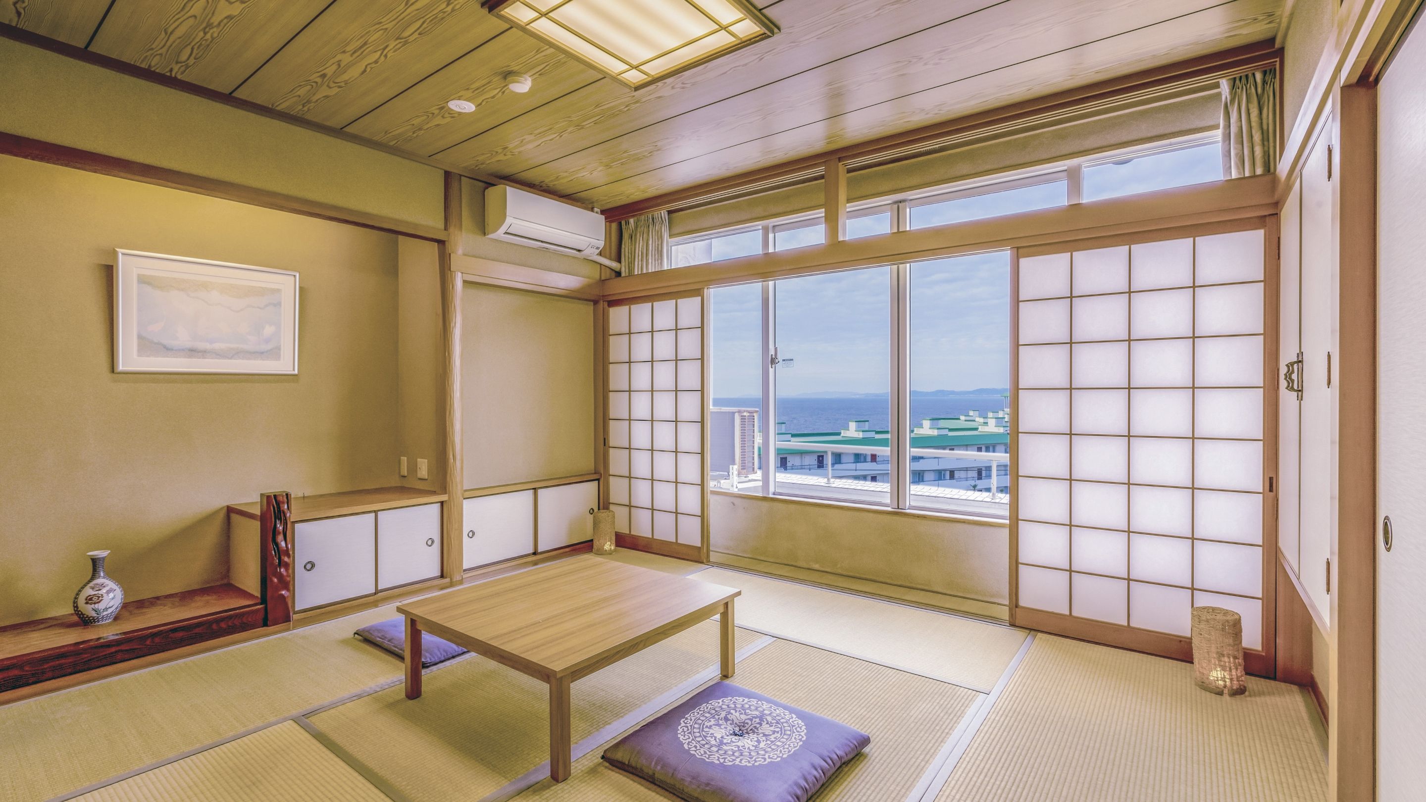 Una habitación japonesa tradicional con suelo de tatami, una mesa de madera baja, un banco acolchado y un amplio celosía de papel con vistas a edificios y al mar.