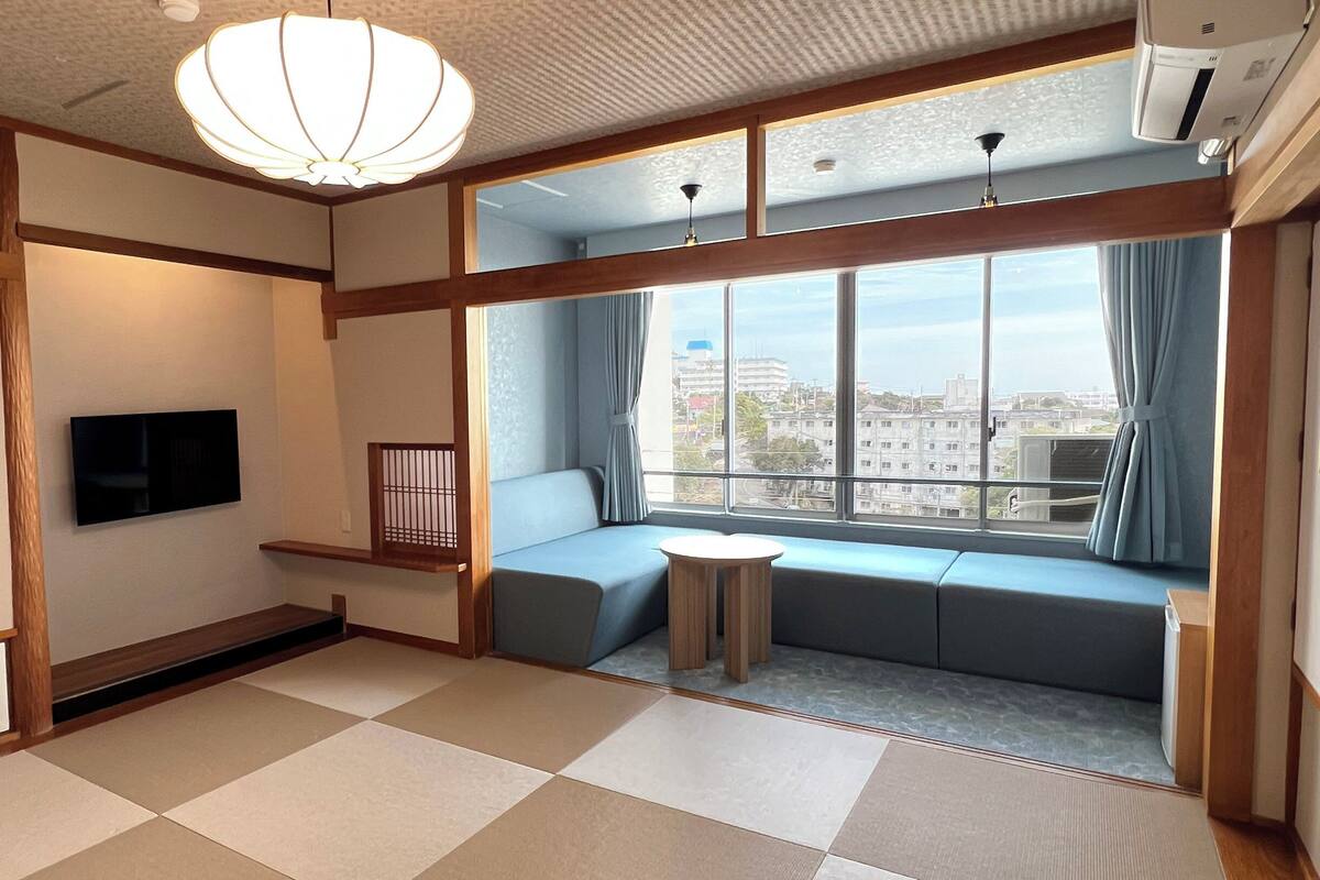 A room with tatami flooring, a low table, a sofa, a TV, and large windows with curtains.