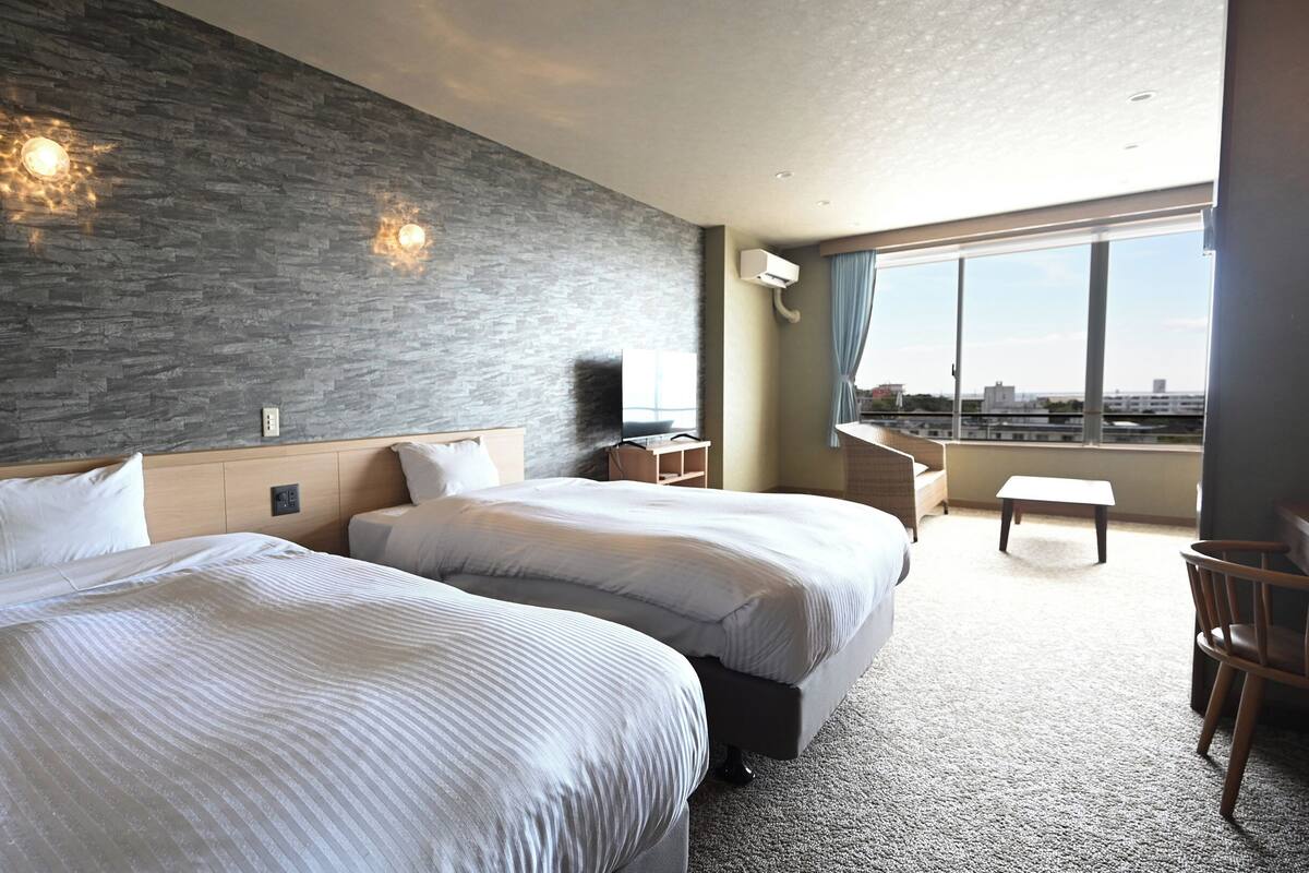 A hotel room with two beds, a TV, a desk, and a view of the city.