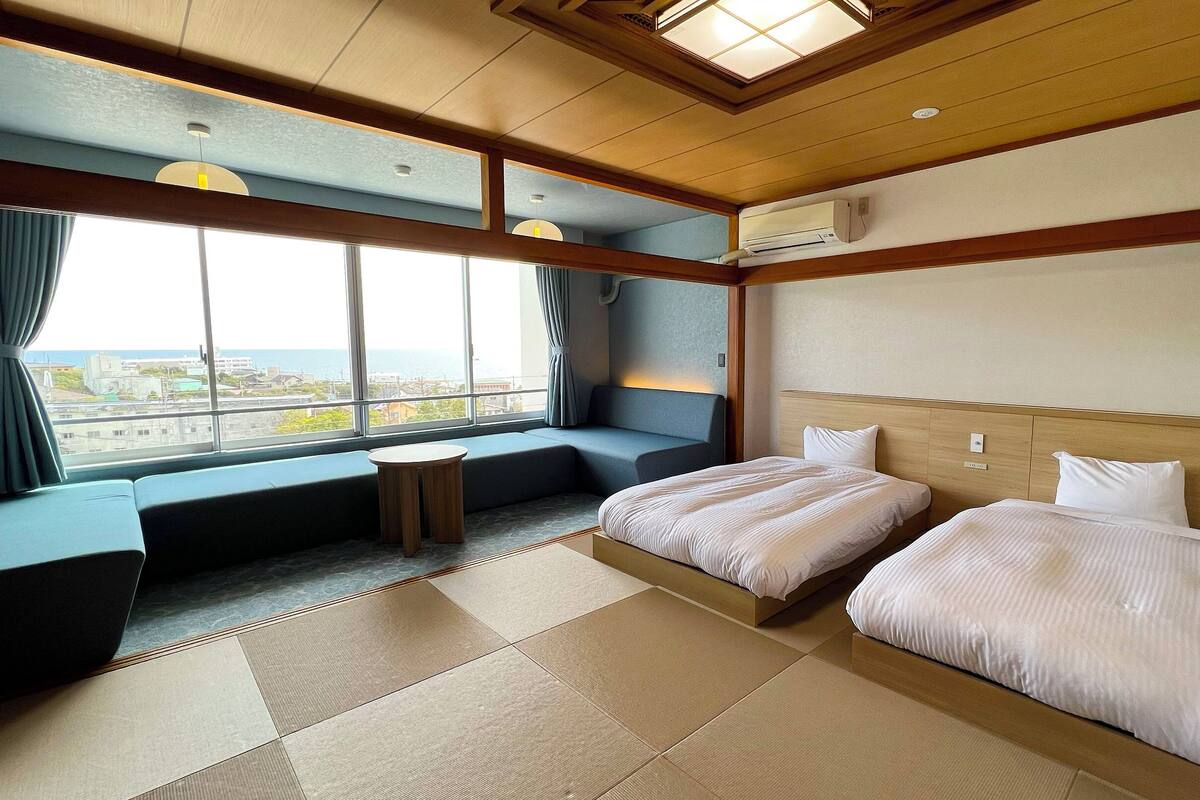 A hotel room with two beds, a seating area, a small table, and a large window offering a view.