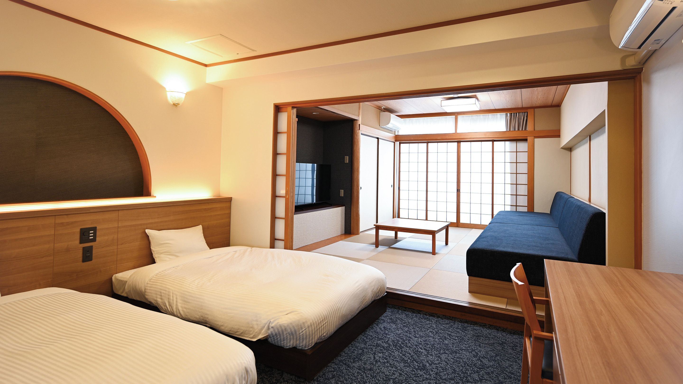 A hotel room with two beds, a sofa, a TV, and a sliding door leading to a balcony.