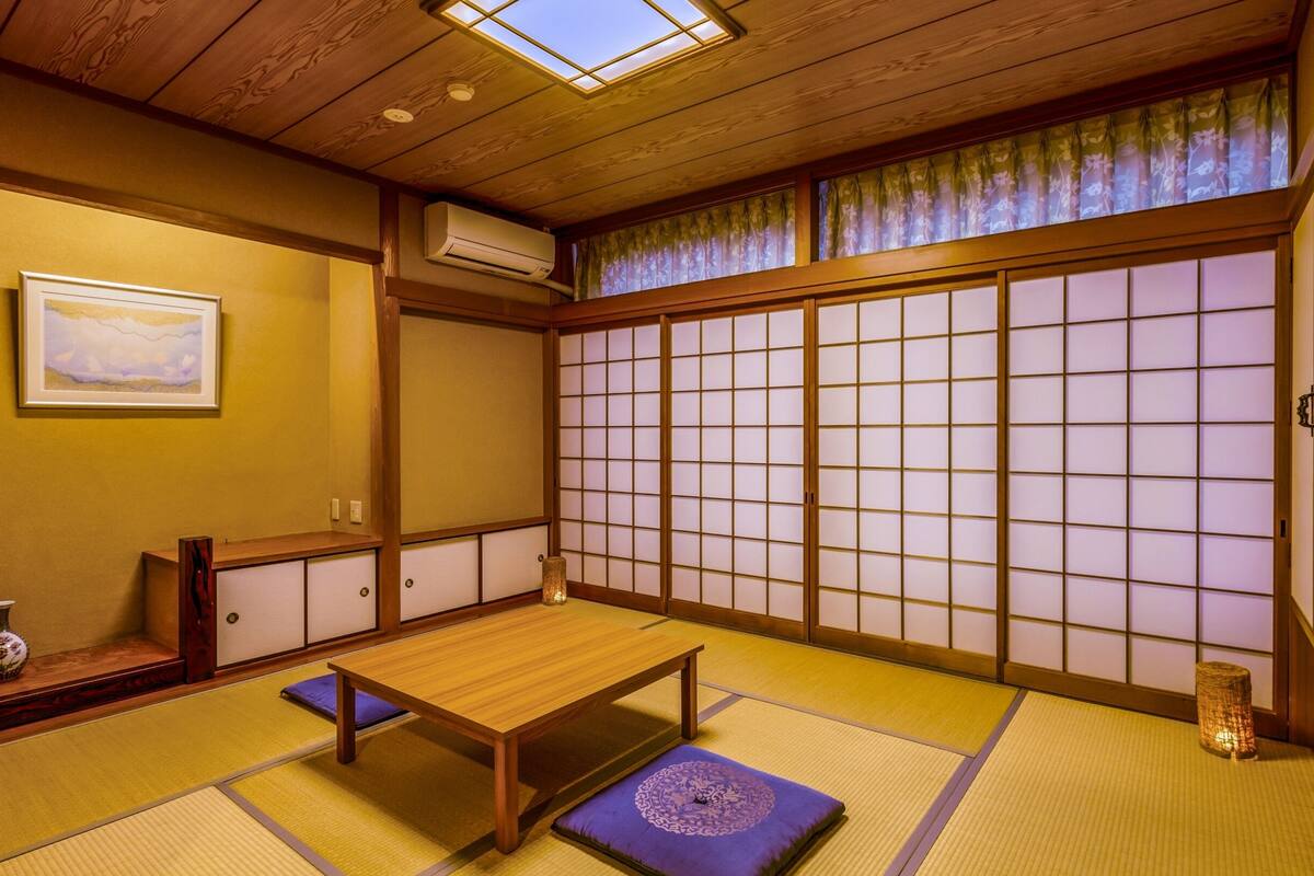 A traditional Japanese room with tatami flooring, a low wooden table, a cushion, a vase, and a painting on the wall.