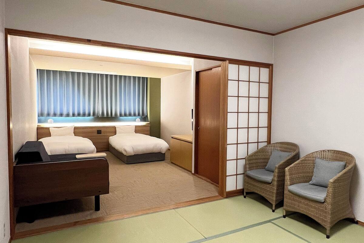 A hotel room with a bed, a desk, two chairs, a sliding door, and a view through a window with curtains.