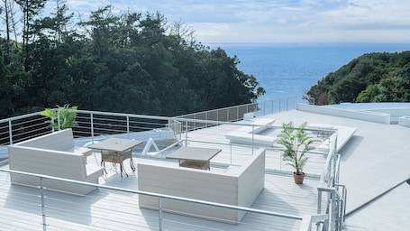 A rooftop terrace with a view of the sea, furnished with tables and chairs, surrounded by greenery.
