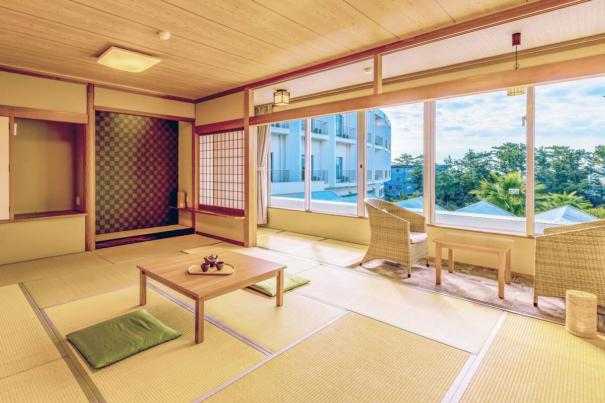 A traditional Japanese room with tatami flooring, a low table with a tray, a green cushion, and a view of a building through large windows.