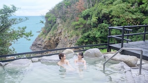 A hot spring with two people in the water, surrounded by rocks and a view of the sea and cliffs.