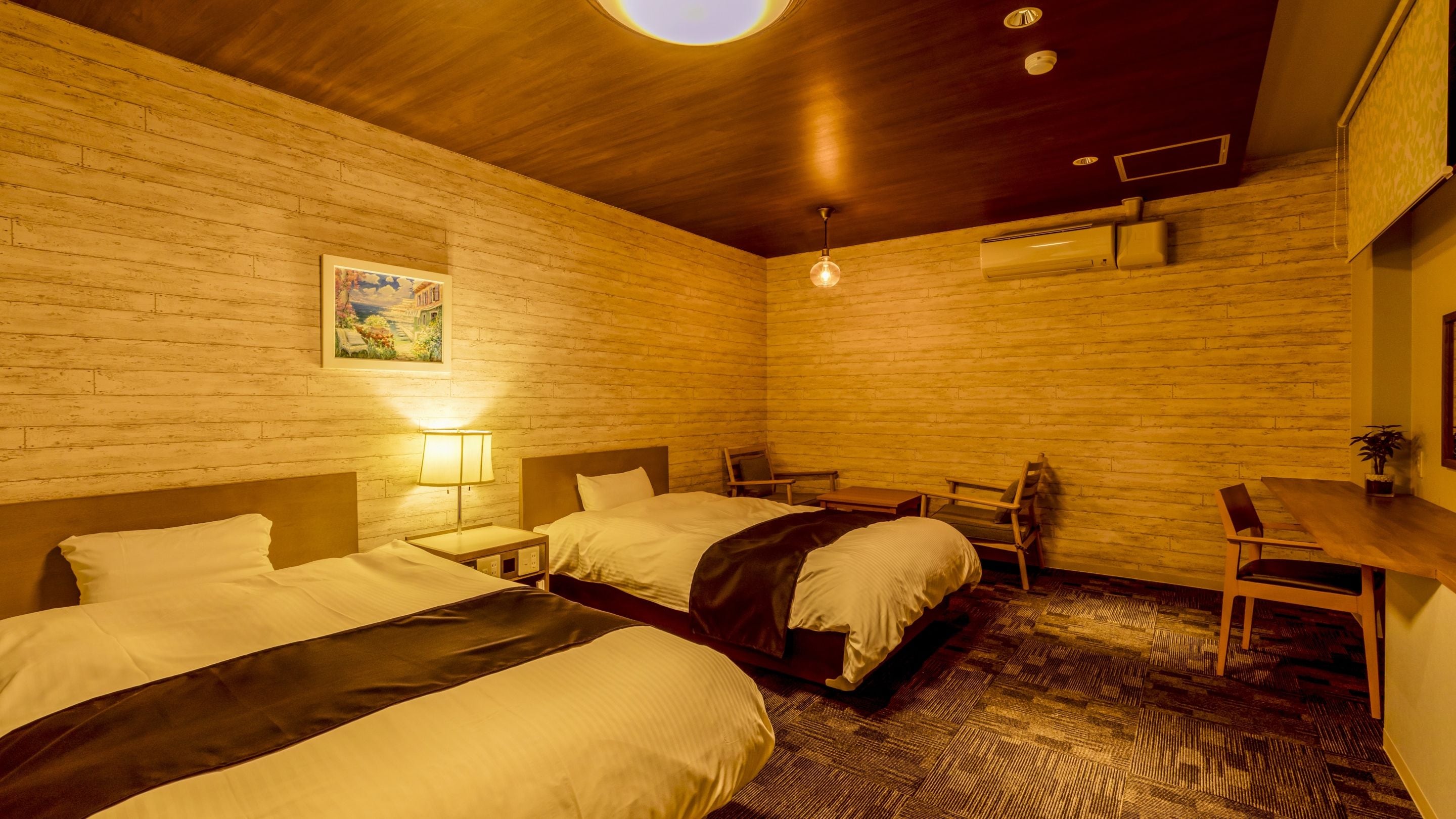 A hotel room with two beds, a desk, a chair, a lamp, and a painting on the wall.