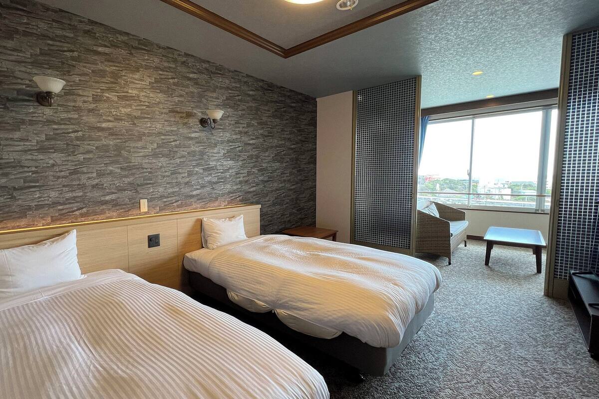 A hotel room with two beds, a stone wall, a large window, and a view of the city.
