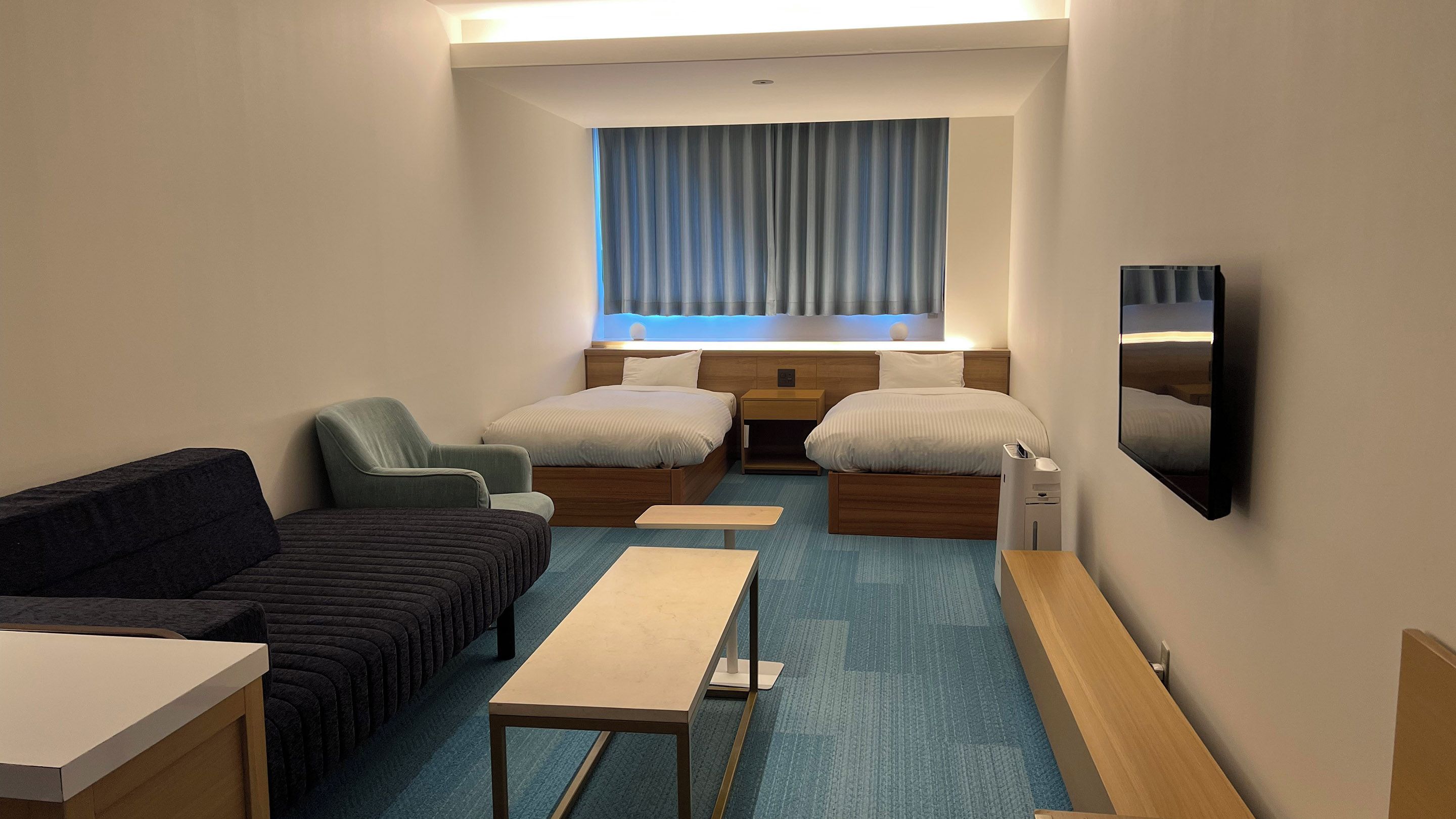 A hotel room with two beds, a sofa, a small table, a TV, and a window with curtains.