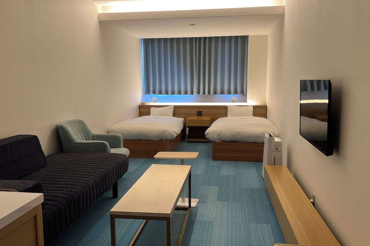 Une chambre d’hôtel avec deux lits, un canapé, une petite table, une télévision et une fenêtre avec des rideaux.