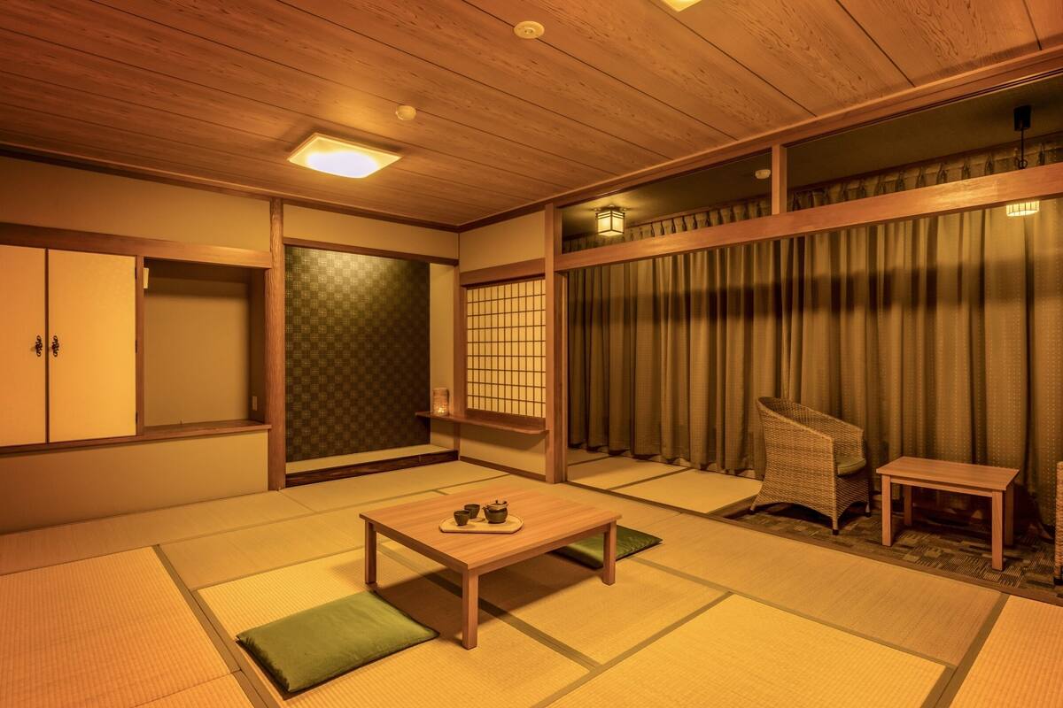 A traditional Japanese room with tatami flooring, a low wooden table, a green cushion, a wicker chair, and a sliding door with a shoji screen.